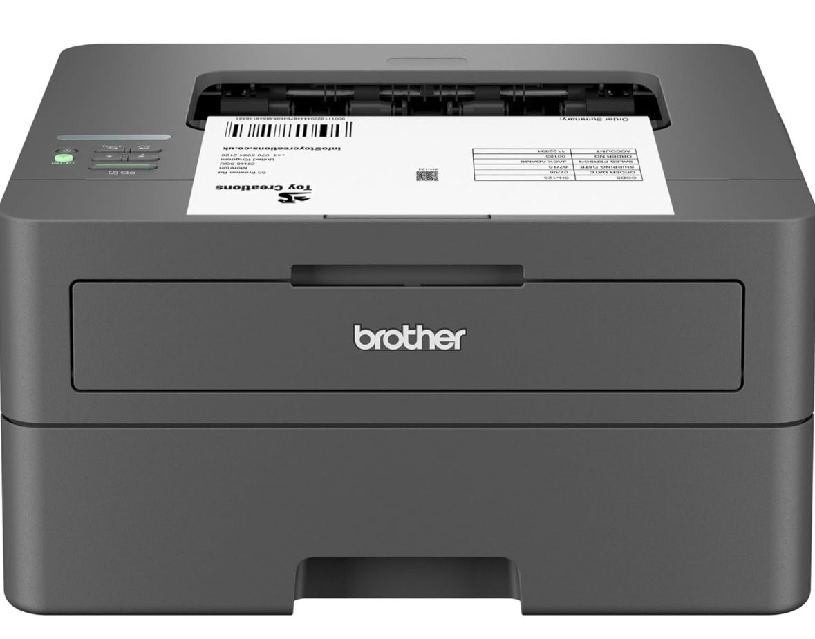 A dark gray Brother laser printer is shown from a slightly elevated front view, with a white sheet of paper partially ejected from the top output tray. The paper displays a barcode and text, including an "Order Summary" and contact information for "Toy Creations."