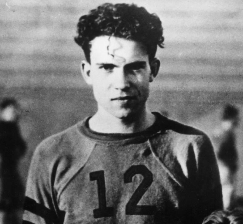 A black and white photo shows a young man with dark, curly hair looking directly at the camera. He is wearing a long-sleeved shirt with the number "12" on the front.