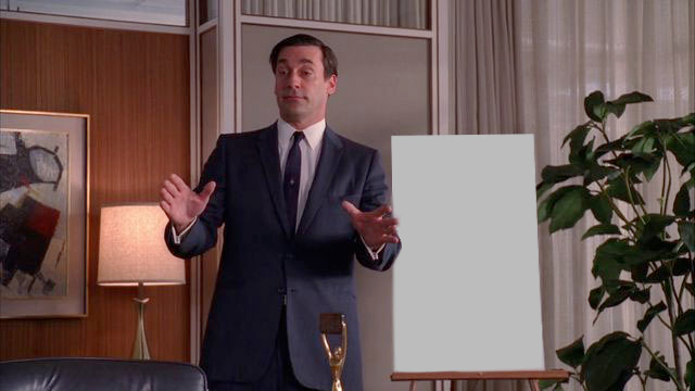A man in a suit stands in an office, gesturing with both hands towards a blank white board on an easel to his right. To his left is a framed abstract painting and a lamp, and behind him are wood-paneled walls and a window with sheer curtains.