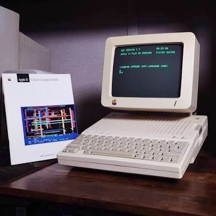 An old Apple II Plus computer with a beige monitor and keyboard sits on a dark wooden desk next to a white book titled "Apple II A Touch of AppleSoft BASIC." The monitor displays green text on a black screen.