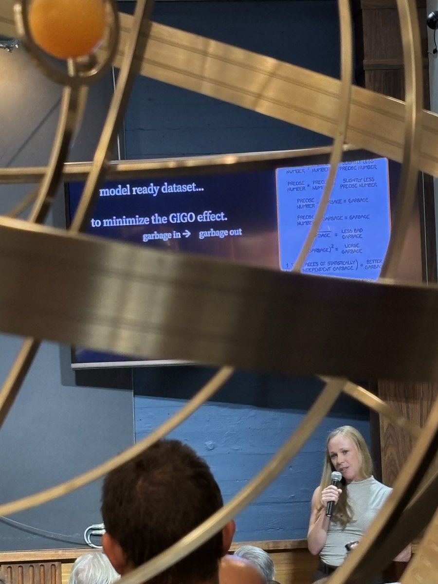 A woman with long blonde hair speaks into a microphone, standing in front of a blue wall. In the background, a screen displays text about "model ready dataset" and "GIGO effect." The foreground is obscured by a large, ornate metal structure.
