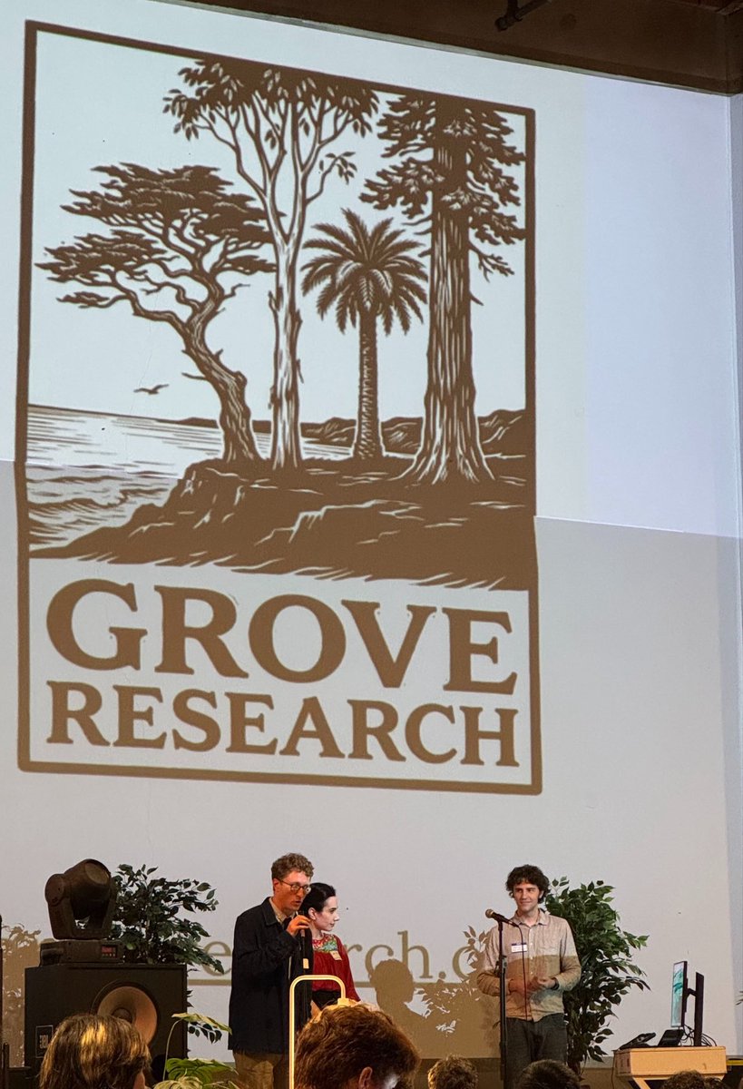 A large screen displays the "Grove Research" logo, featuring a stylized illustration of various trees on a coastline with the ocean in the background. Below the screen, three people are on a stage, with two men speaking into microphones and a woman standing between them.