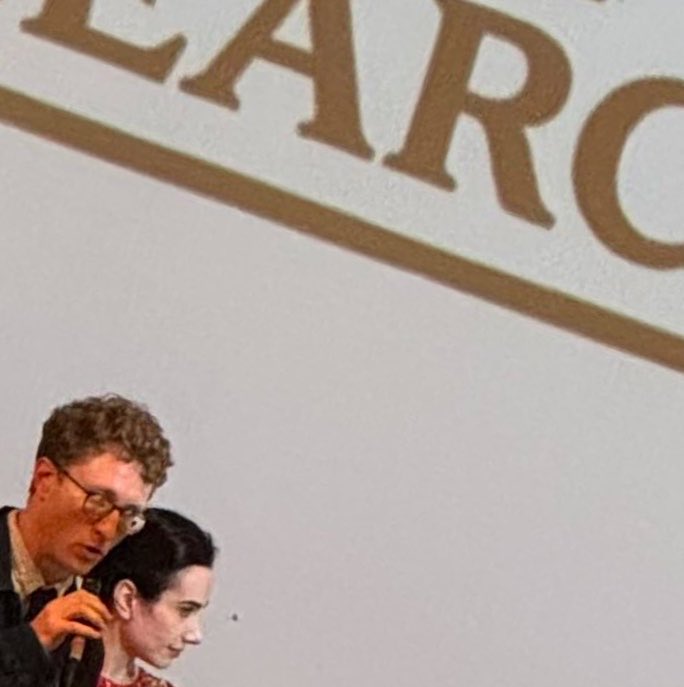A man with curly hair and glasses holds a microphone near a woman with dark hair, both looking to the right. Above them, large golden letters are visible on a white background.