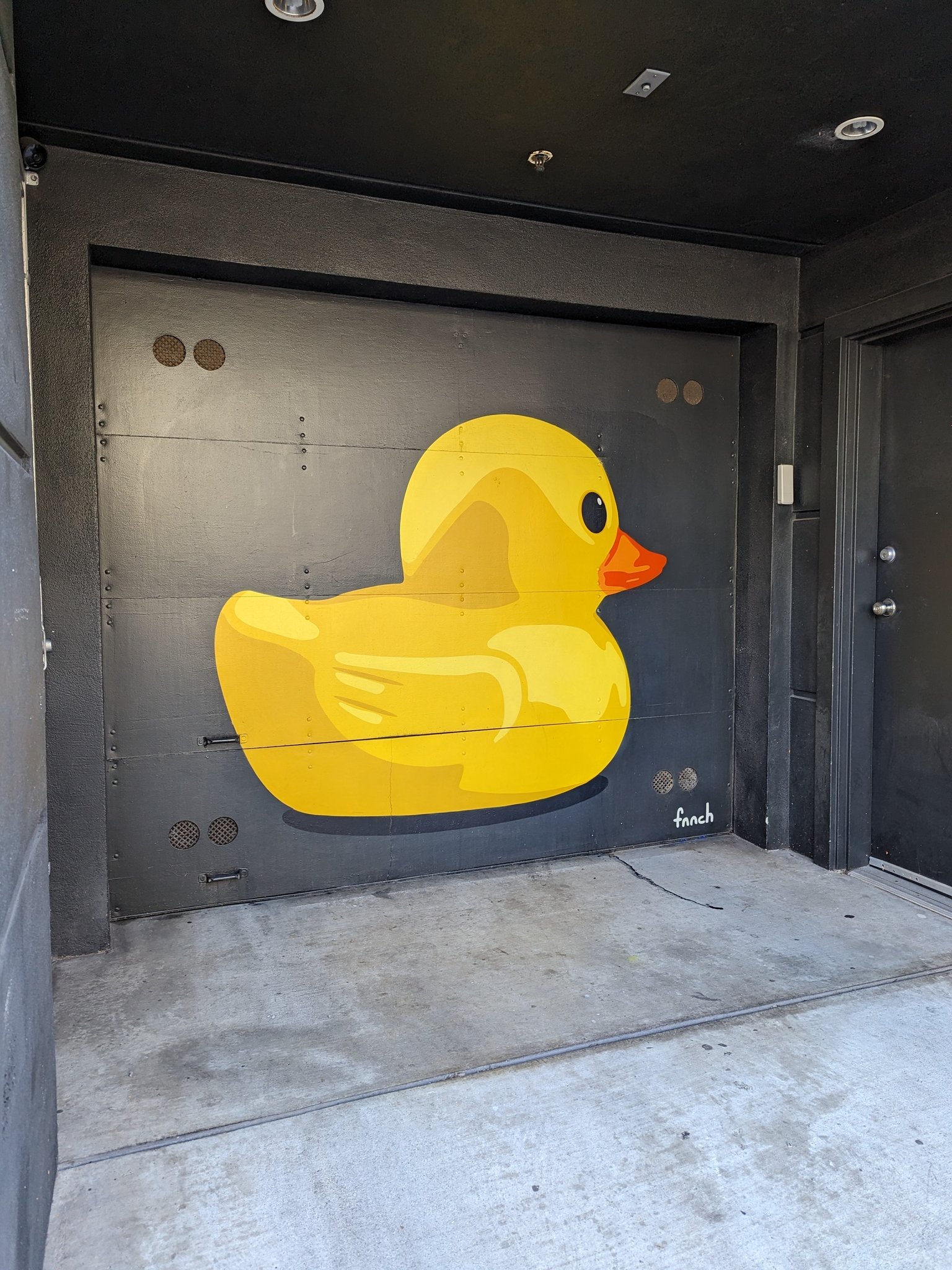 A large mural of a yellow rubber duck with an orange beak is painted on a black garage door. The artist's signature, "fnnch," is visible in the bottom right corner of the door.