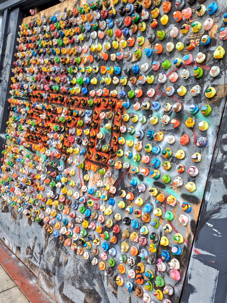 Hundreds of small, colorful rubber ducks are mounted in a dense grid pattern on a large, weathered wooden board. The board is covered in layers of gray, black, and orange spray paint, creating a textured, urban backdrop for the diverse collection of ducks.