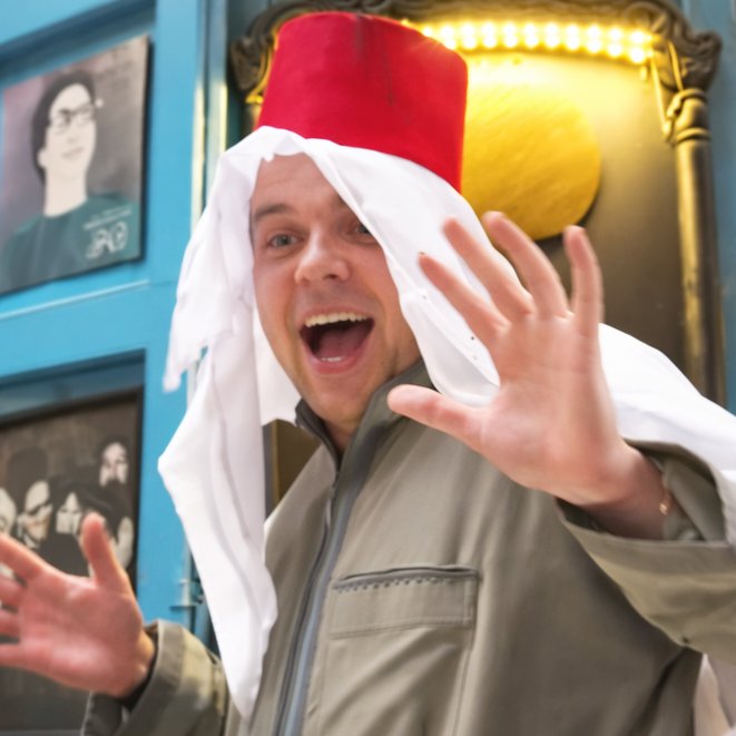 A man with light skin and short brown hair smiles with his mouth open, wearing a red fez and a white ghutra over a light green thobe. He holds his hands up with palms facing forward, as if in surprise or greeting.