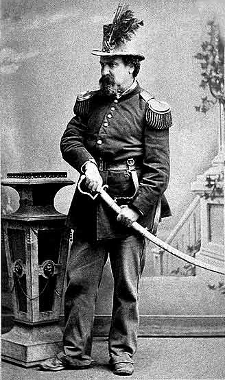 A full-length black and white studio portrait of a man in a military-style uniform with epaulettes and a feathered hat, holding a sword in its scabbard. He stands next to an ornate pedestal in front of a painted backdrop.