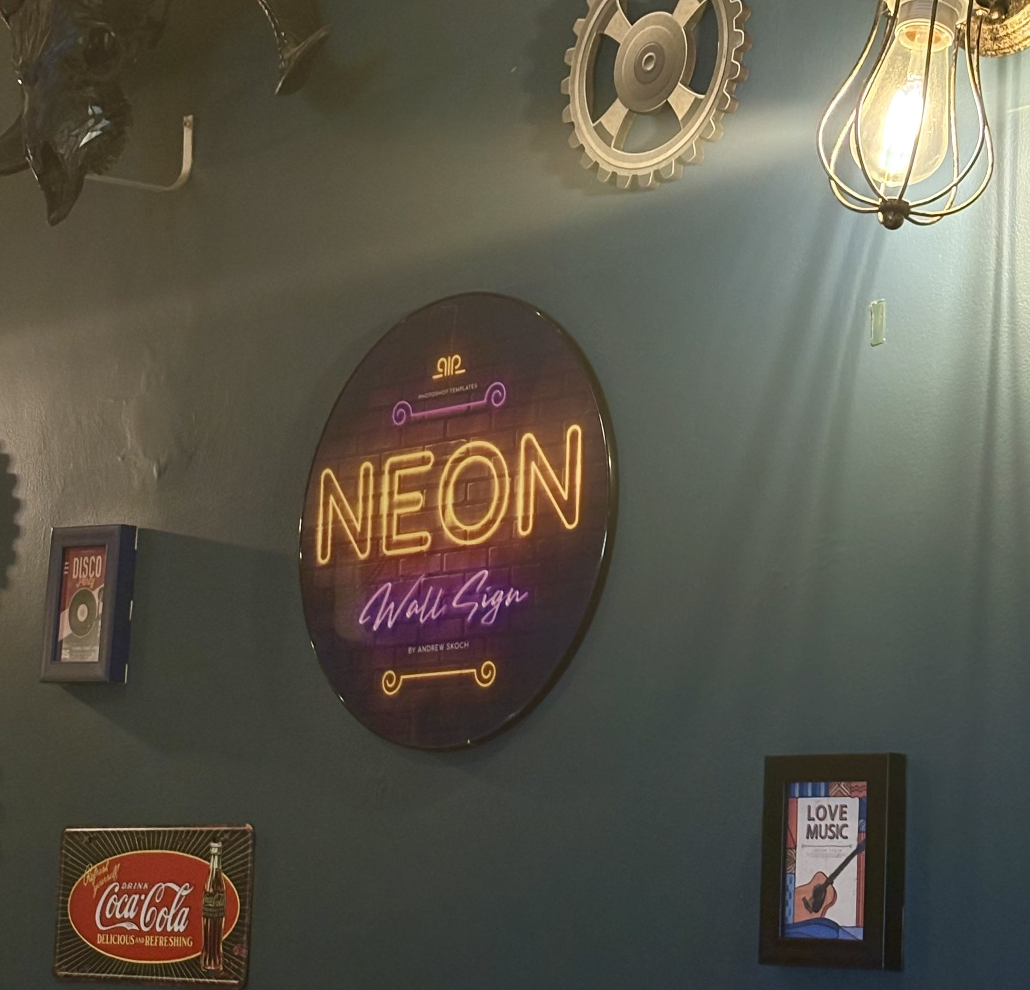 A dark blue wall is decorated with various items, including a large circular sign with glowing neon text, a framed "Disco Party" poster, a Coca-Cola tin sign, and a framed "Love Music" poster. A light fixture with a caged bulb illuminates the upper right corner.