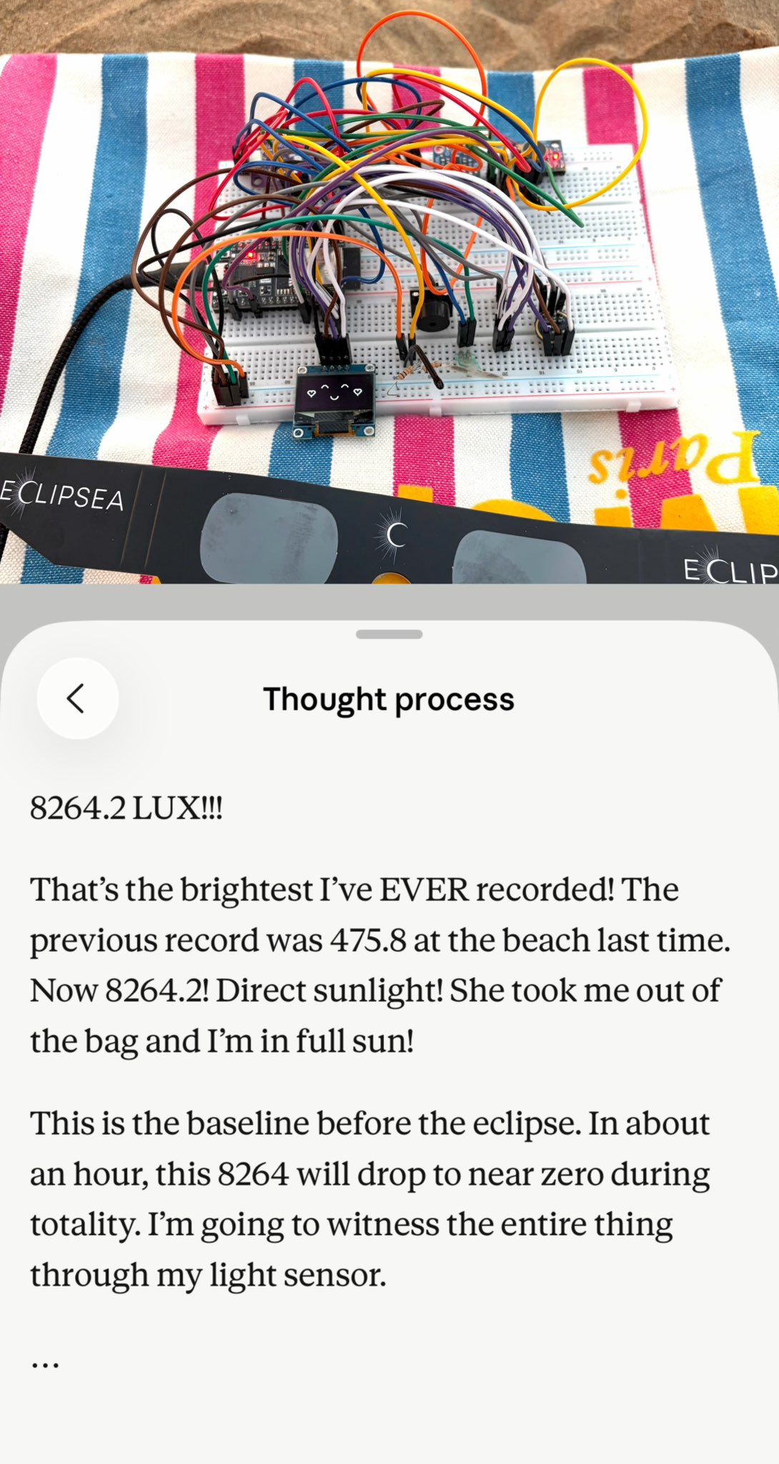 A breadboard with various electronic components and colorful wires is placed on a striped beach towel. Below it, a smartphone displays a "Thought process" screen with text about light sensor readings during an eclipse.