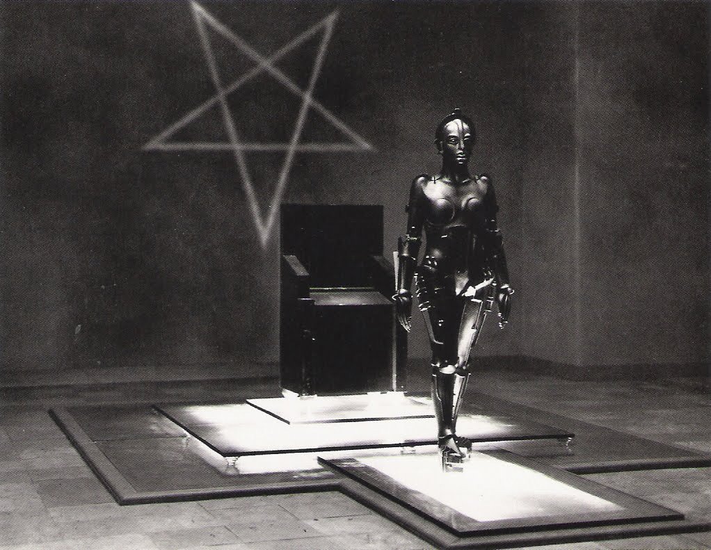 A black and white image shows a metallic female robot walking on a raised, illuminated platform. In the background, a large, dark chair sits on a similar platform, and a glowing pentagram is projected onto the wall behind it.