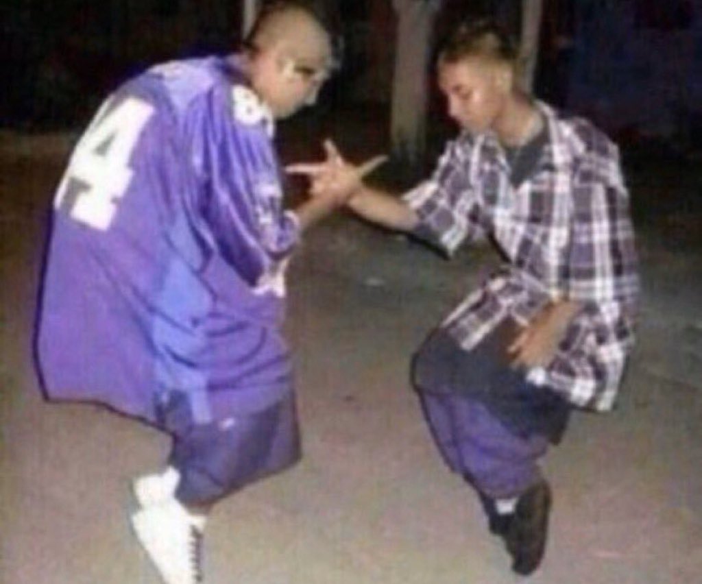Two men are crouched down, facing each other, with their hands clasped together in a handshake. The man on the left wears a purple jersey with the number "4" on it, while the man on the right wears a purple and white plaid shirt.