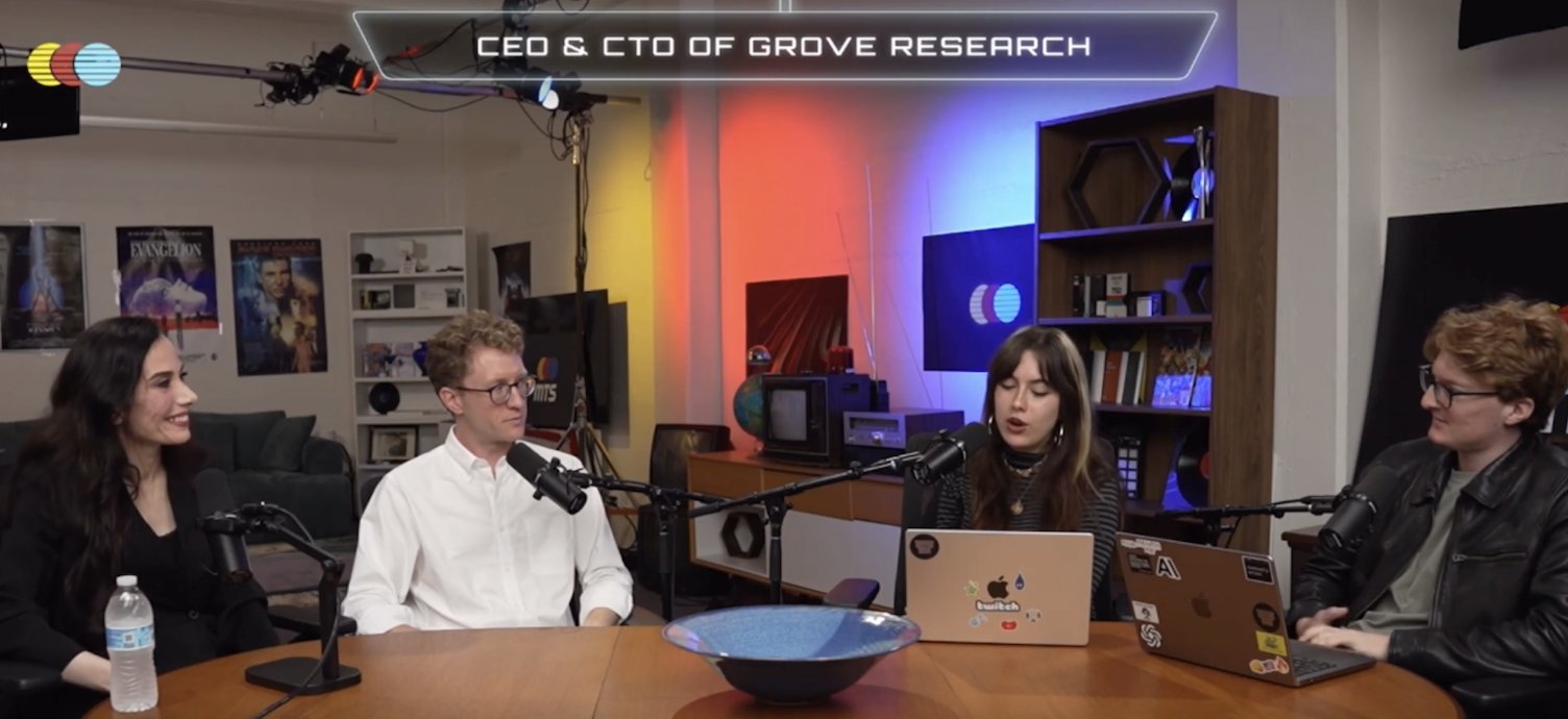 Four people, two men and two women, are seated around a wooden table with microphones in front of them, participating in a podcast or interview. The background features shelves with various items, movie posters, and colorful lighting.