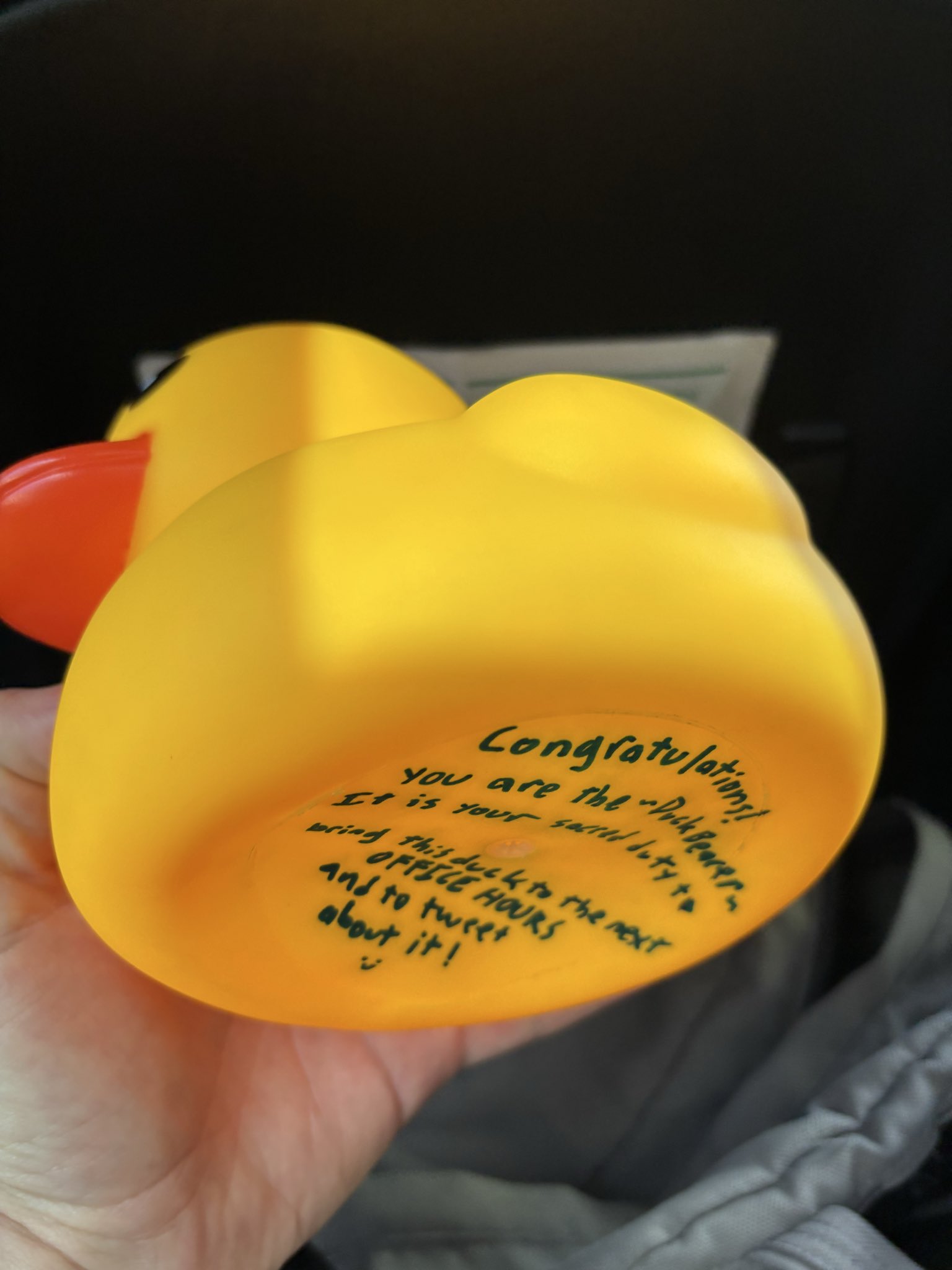 A person holds a yellow rubber duck with an orange beak. Black handwritten text is visible on the bottom of the duck.