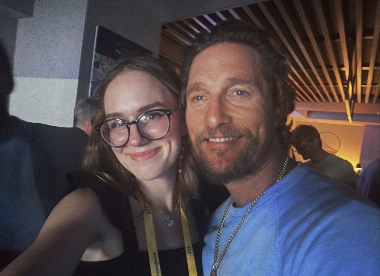 A young woman with glasses and a man with a beard are smiling at the camera in a selfie. The woman is on the left, wearing a black top and a yellow lanyard, and the man is on the right, wearing a light blue shirt and a silver chain.