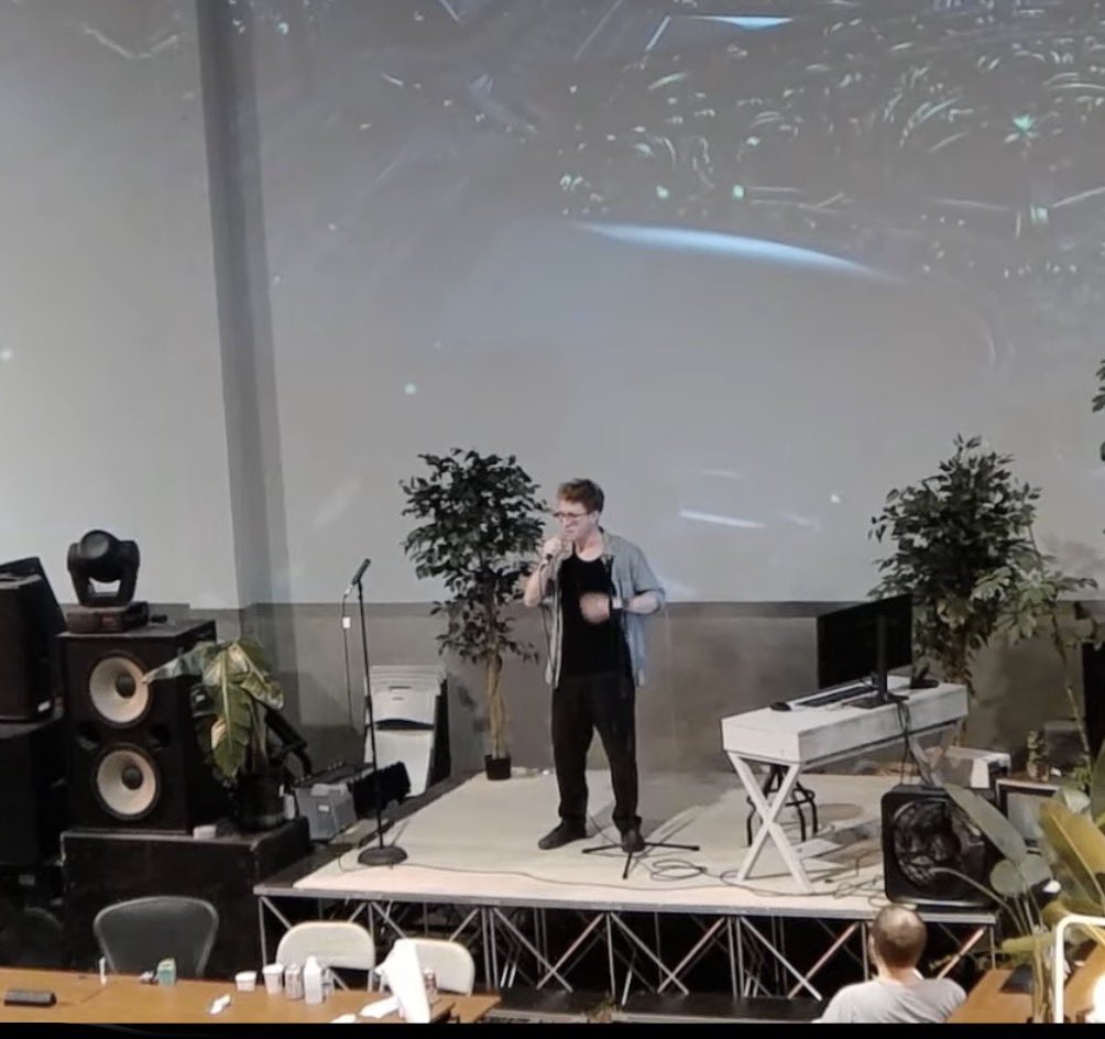 A man with glasses and a light blue shirt over a black t-shirt stands on a stage, holding a microphone and singing. Behind him is a large screen displaying an abstract image, and on either side of him are potted plants.