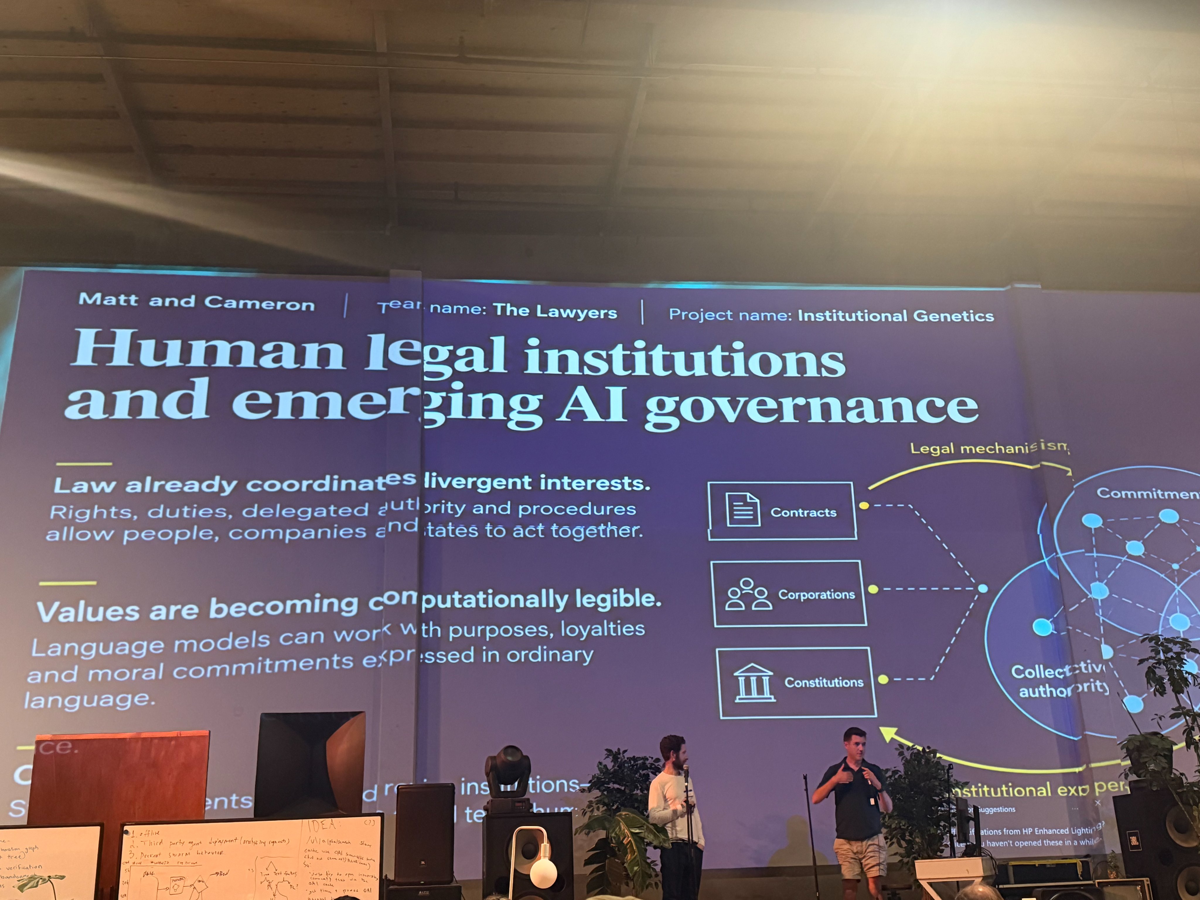 Two men stand on a stage in front of a large screen displaying a presentation titled "Human legal institutions and emerging AI governance." The man on the left has a beard and is wearing a light-colored long-sleeved shirt and dark pants, holding a microphone. The man on the right is wearing a dark polo shirt and light-colored shorts, gesturing with his hands while holding a microphone.