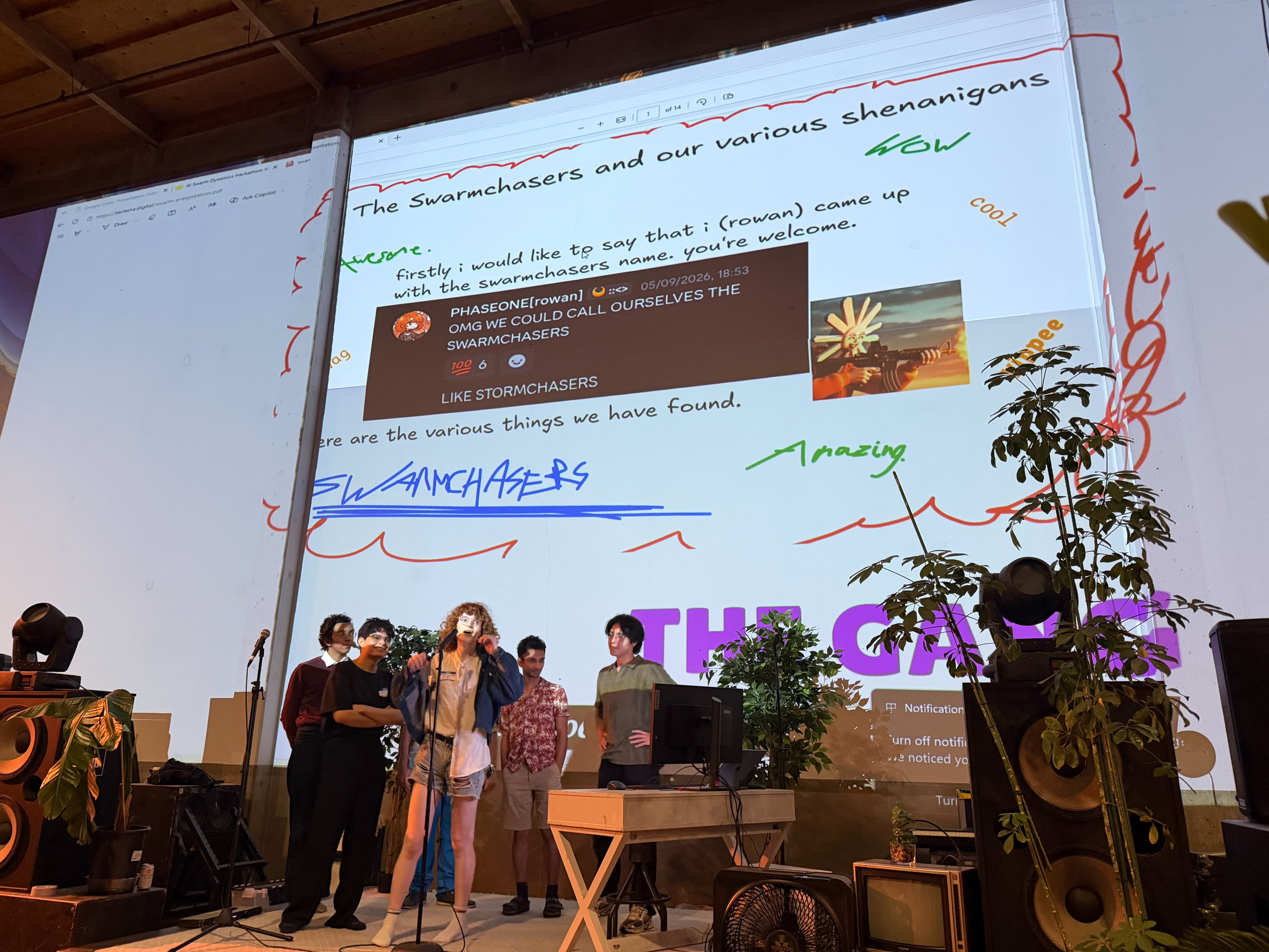 A group of young adults stands in front of a large screen displaying a presentation with text and images. The person in the center is speaking into a microphone, wearing a denim jacket and ripped shorts.