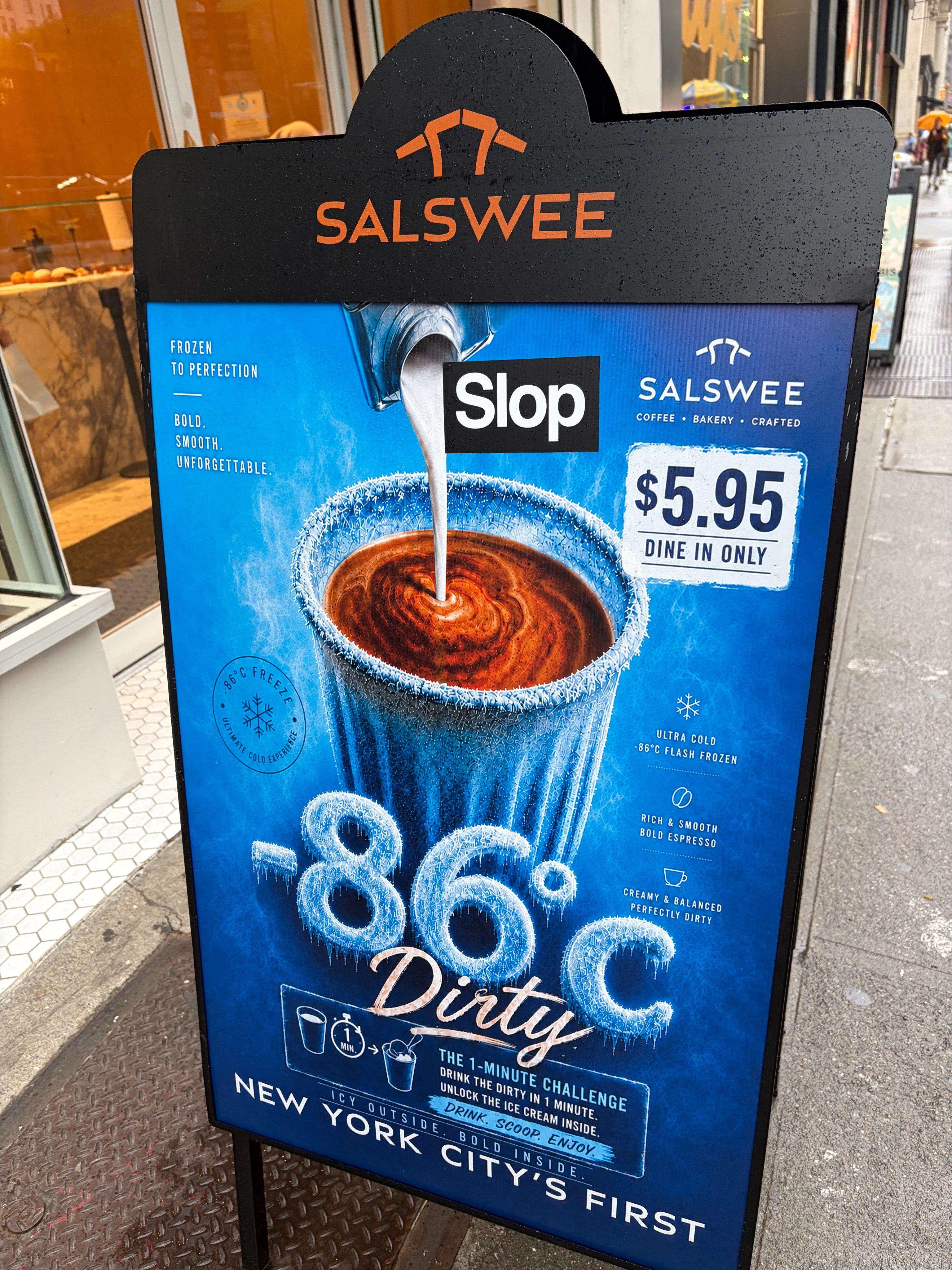 A black A-frame sign with a blue background advertises a frozen drink called "Slop" or "Dirty" from Salswee. The sign shows a white liquid being poured into a frosty cup with a brown, swirled drink, and text describes the drink as "frozen to perfection" and "ultra cold -86°C flash frozen."